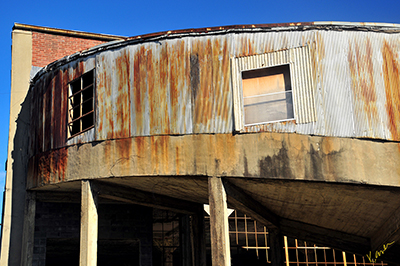 Photo of an old abandoned factory with curved walkways and busted out windows, red rusty metal and brick against a blue sky